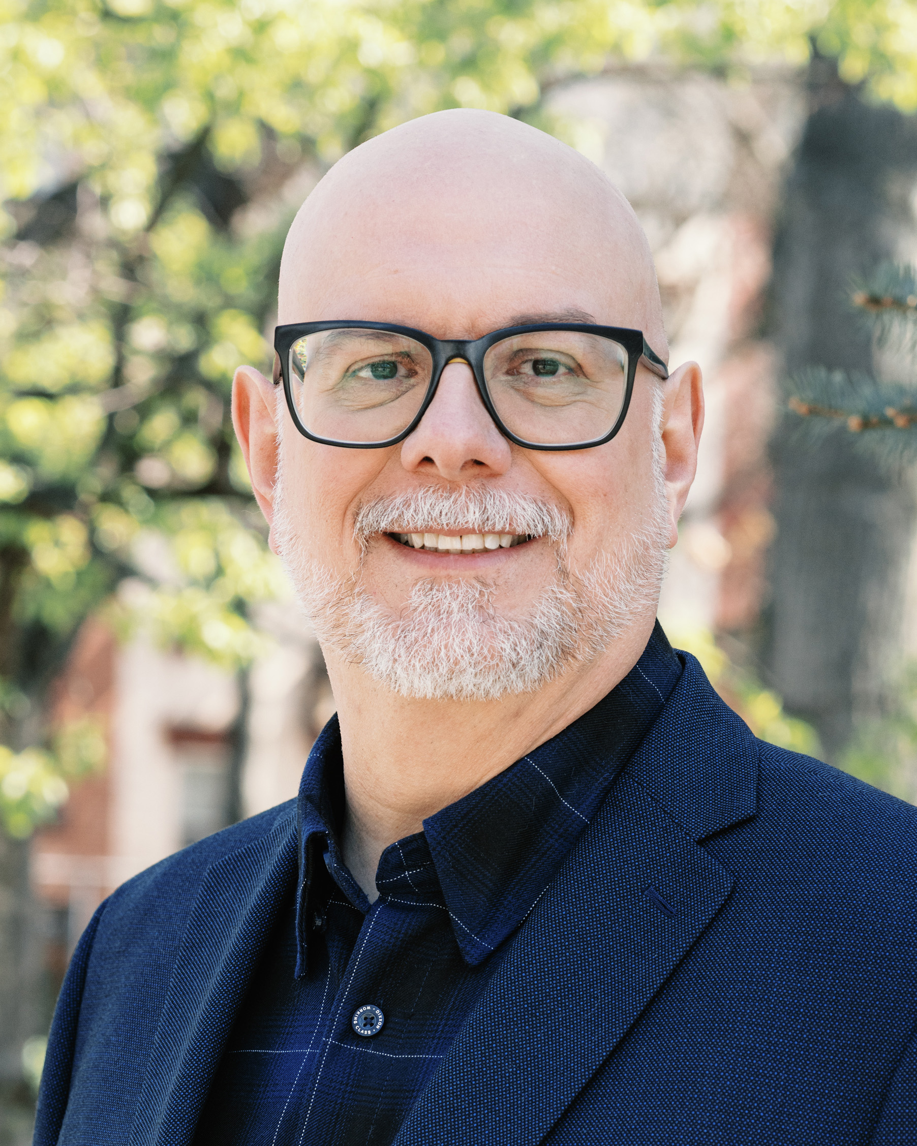 Smiling bald man with glasses and a white beard, wearing a dark blue blazer and checked shirt outdoors.